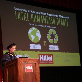 Scholar Ben Callard stands at a podium in front of a screen announcing the 2019 Latke Hamantash Debate at the University of Chicago