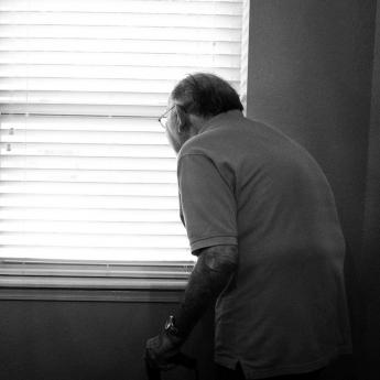 Elderly man standing with cane looking out window
