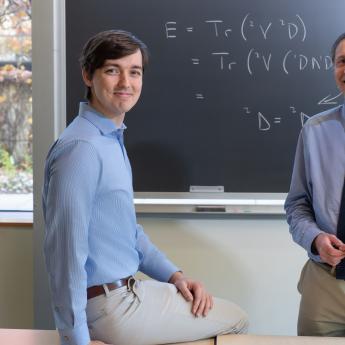 two scientists in blue button-down shirts smiling at camera in front of a chalkboard with equations