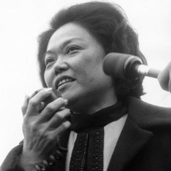 Black and white photo of Rep. Mink in a suit speaking into a handheld microphone, looking to the side