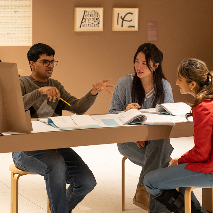 Three students engaged in discussion while sitting at a table. Exhibit artwork is visible behind them.