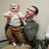 Smiling baby, newly outfitted with cochlear implant, held by his smiling father as mother looks on.