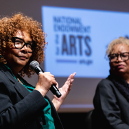 Julie Dash sits on stage while holding and talking into a microphone. Next to her on the right sits Barbara McCullough. 