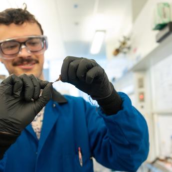 Photo of A scientist with black gloves and blue lab coat and a mustache holding a small item