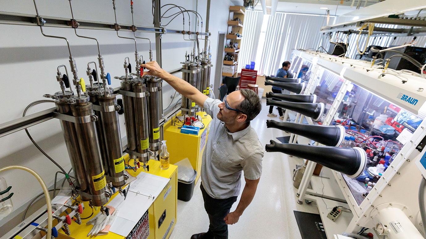 Scientist in goggles reaches up to turn knobs on a row of tubes in a laboratory