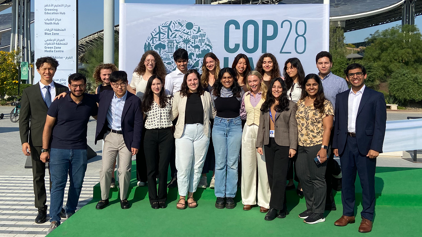 A group of 18 people standing in two rows pose in front of a sign that says COP28