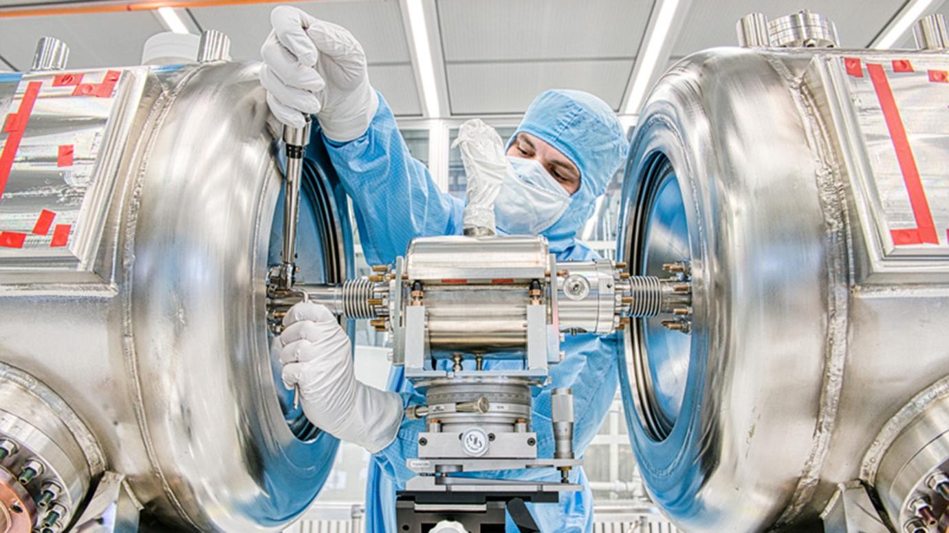 Technician in full-body blue suit, gloves, and mask leans in with a tool between two large silver cylinders