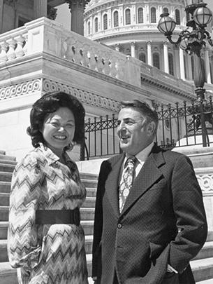 Black and white photograph of two people dressed in suits outdoors on the steps of the Capitol on a sunny day, wearing suits. Mink is smiling with her face turned to the camera; Mikva is smiling and looking to the left