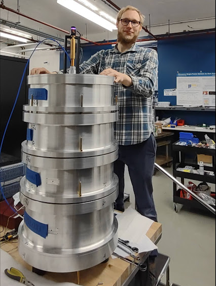 Photo of a scientist in plaid shirt smiling and standing next to a tall metal cylinder with wires and pliers scattered around