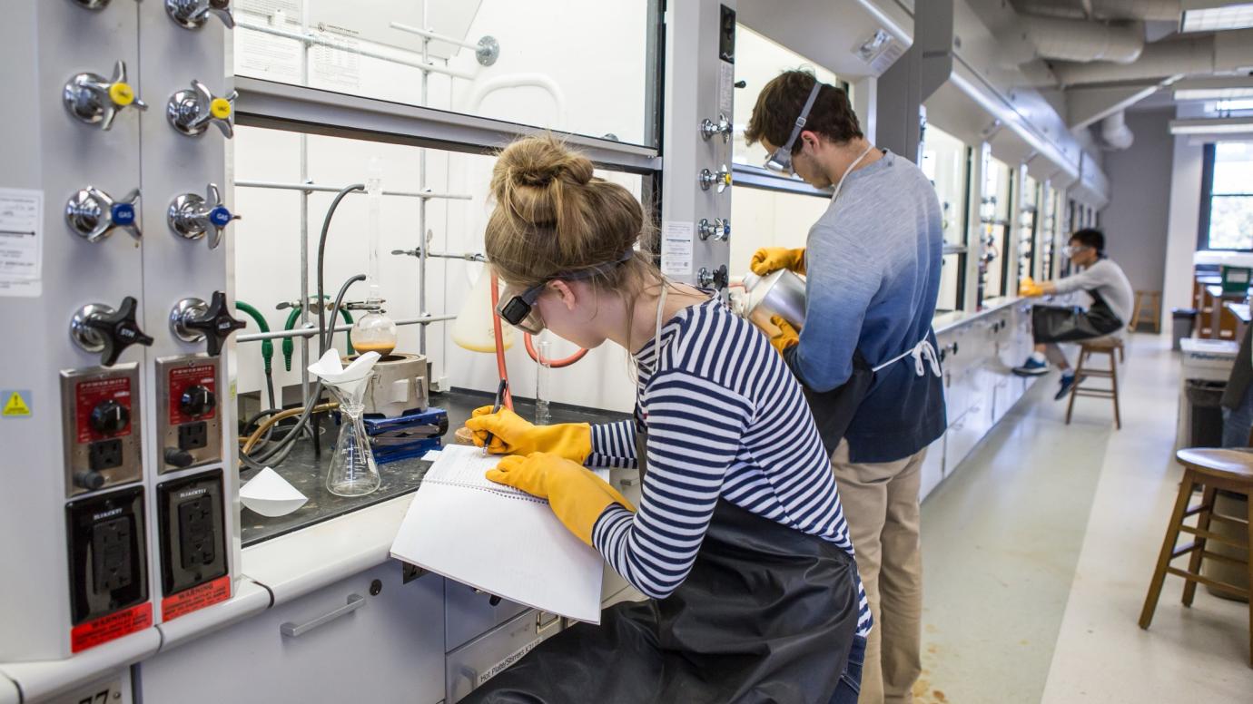 Photo of several student researchers workin in a lab at a fume hood, wearing gloves and safety goggles, one writing in a notebook, with their backs turned to the camera