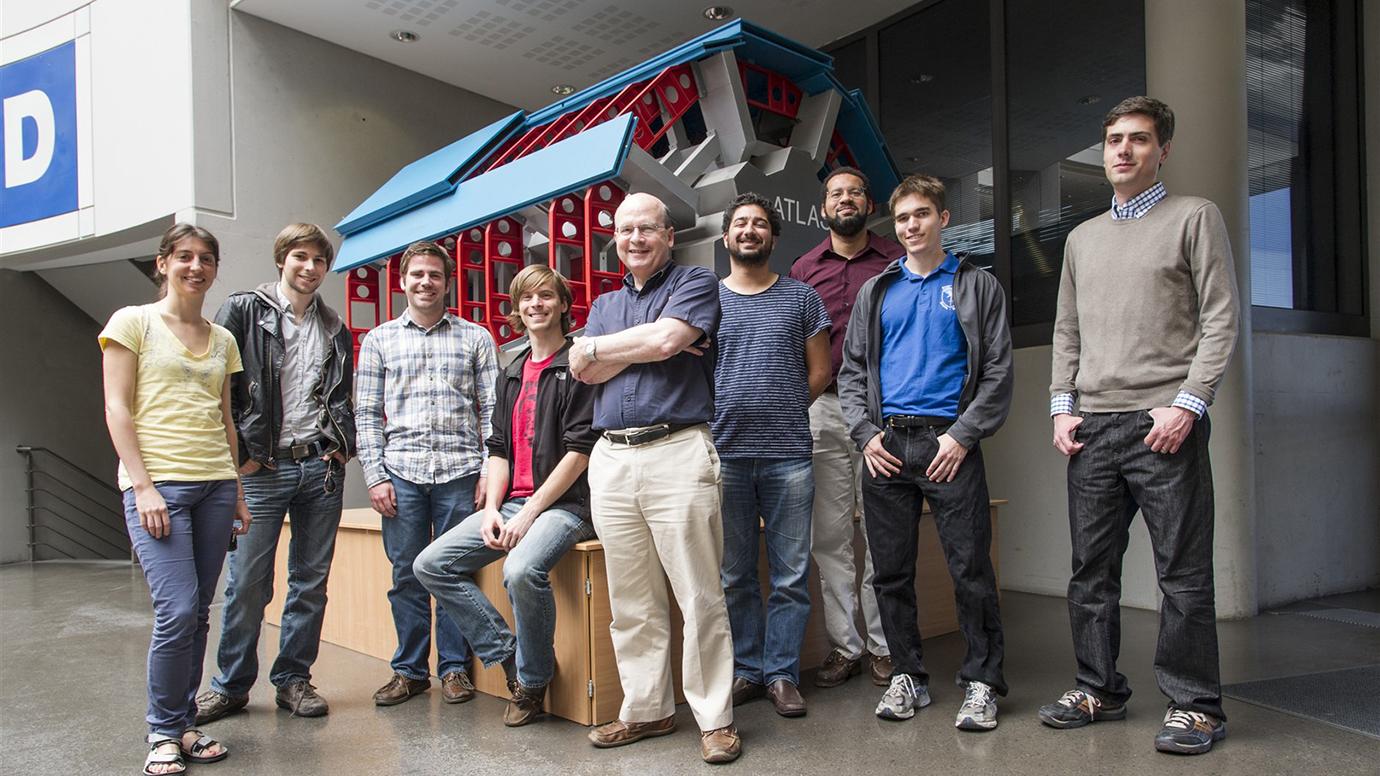 Group of researchers stands in a CERN lobby in front of a model of part of the accelerator