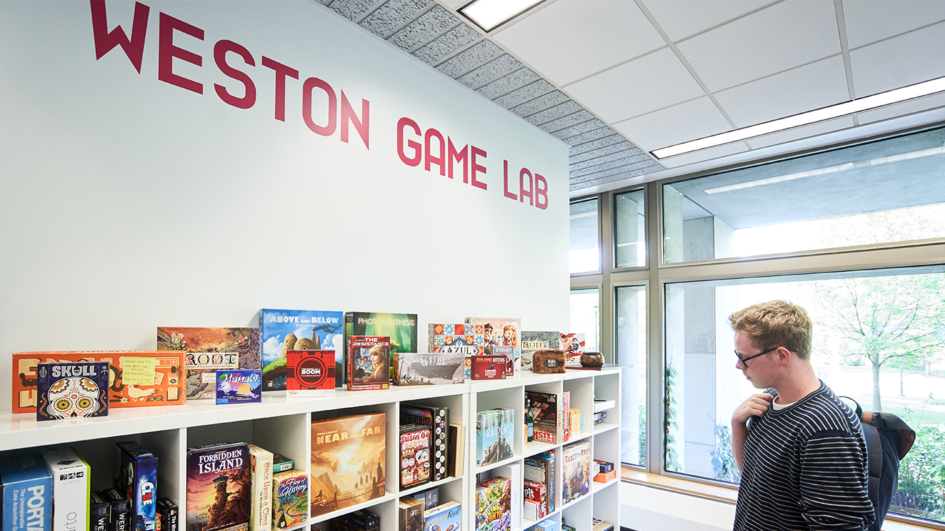 A person stands near shelves filled with board games in a bright room labeled "Weston Game Lab" with large windows overlooking greenery.