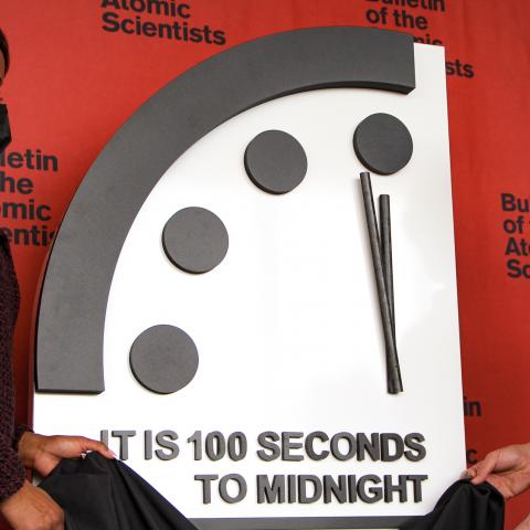Two scholars in masks pull the cloth from a large clock reading "it is 100 seconds to midnight"