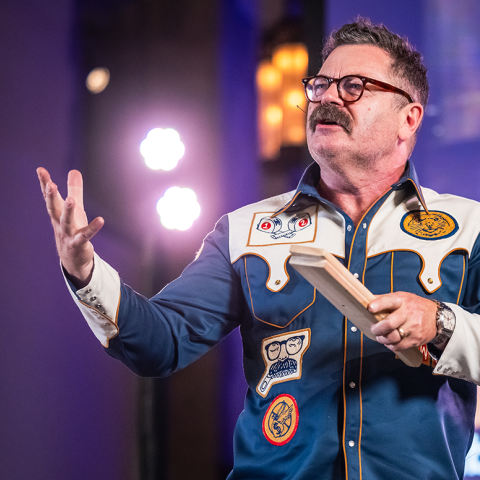 Nick Offerman in a colorful shirt with patches speaks on stage, holding a wooden block with bright lights in the background.