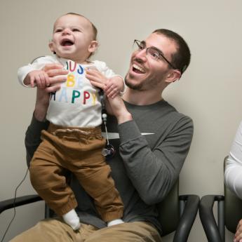 Smiling baby, newly outfitted with cochlear implant, held by his smiling father as mother looks on.