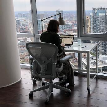 Sarah Cobey working at her desk in a high-rise apartment overlooking Chicago, with dog curled up in a bed next to her