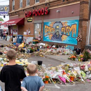 A crowd gathers before a mural of George Floyd at Cup Foods in Minneapolis, with flowers on the sidewalk and street.