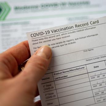 A person holds a COVID-19 vaccine card above an out-of-focus document