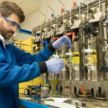 Chemist in a blue lab coat and goggles works with beakers in front of machinery