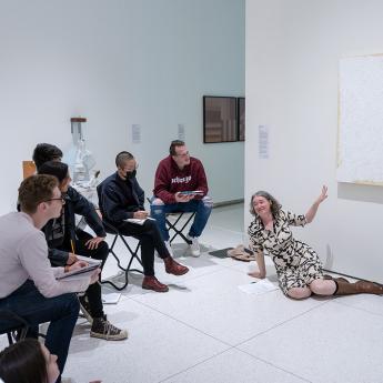 A group of students sit in an art exhibition gallery. They all look at Christine Mehring who is sitting on the floor and pointing up at a white painting by Robert Ryman. 