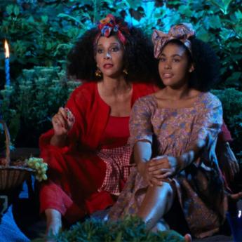 Two black women sit next to each other looking into the distance. The older women in red has her arm around the younger one and is holding a grape. They are surrounded by vegetation and candles. A fruit basket is visible in the foreground.