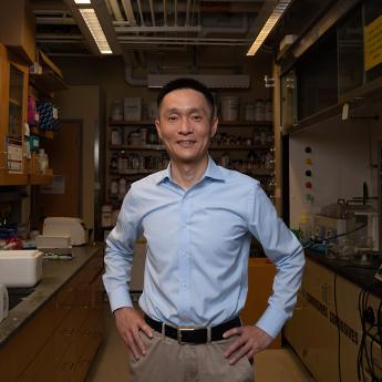 Prof. Chuan He faces camera smiling with hands on hips with a chemistry lab in the background