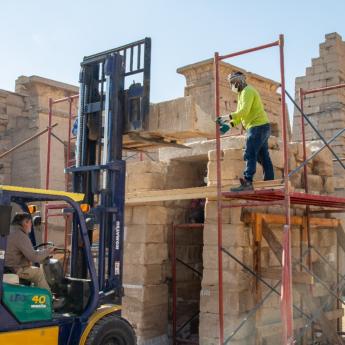 A man standing on scaffolding places a large stone block on a forklift driving by another man. The scaffolding is against a stone gateway. A larger temple complex is visible in the background.