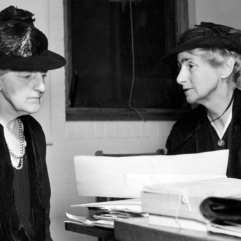 Two women, Sophonisba Breckinridge (right) and Edith Abbott (left) dressed in black and wearing black hats sit next to each other at a desk while examining a document. 