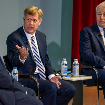 Three men wearing suits sit on stage. The center man, Michael McFaul, gestures while speaking to Robert Pape to the left. Chuck Hagel sits listening on the right.