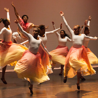 A group of dancers are mid-pose with outstretched arms as they perform onstage in brightly colored skirts.