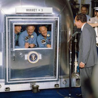 President Nixon stands before three astronauts dressed in blue and contained within a quarantine area