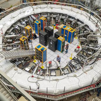An overhead view of a very large round particle accelerator in a building with people working in the center