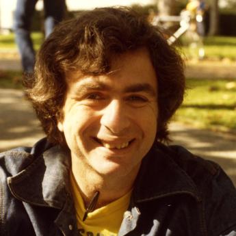 Melvin Rothenberg sitting on ground smiling at camera in a jacket and yellow t-shirt with a pen tucked into it