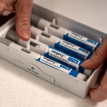 A person reaches from out of frame toward a box of four blue and white syringes, each labeled "Wegovy"