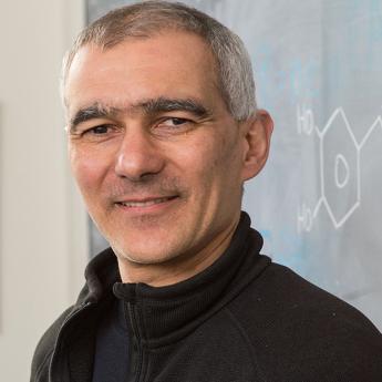 Portrait of Moungi Bawendi looking at camera and smiling in front of a chalkboard with a molecule diagram and a wall photo of beakers containing quantum dots in different colors