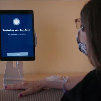 A patient with an IV wearing a mask looks at a tablet screen that reads: "Contacting your care team"