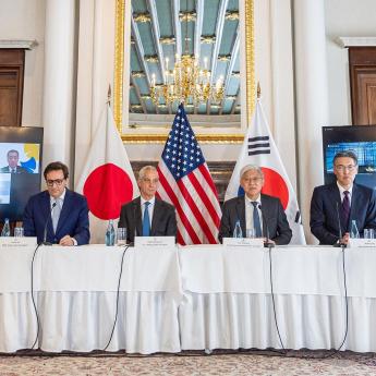 Four men sit at a table covered in a white tablecloth with flags at their back (from left to right: the Japanese flag, the American flag, and the South Korean flag)