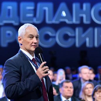 Andrei R. Belousov, a man with short white hair in a suit, holds a microphone as he speaks before an audience