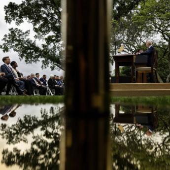 President Biden stands at a lectern delivering a speech to a seated, outdoor crowd. The image is divided in two vertically by a post, and horizontally by a body of water..