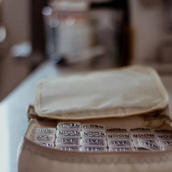 A bag holds a segmented plastic container for pills with each segment variously marked with "NOON," "EVE," and "BED"