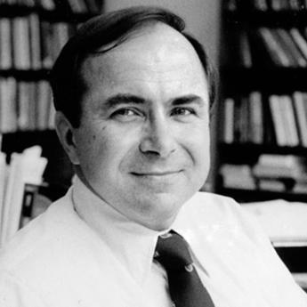 Black and white photo of Mazenko in a white shirt and tie looking at camera in front of bookcases