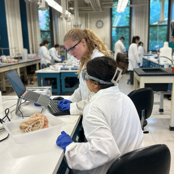 Students in lab coats, gloves and goggles look at a computer sitting on a table with lab equipment.