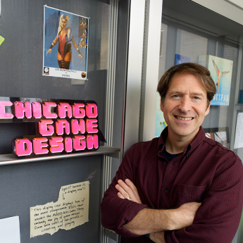 Alex Seropian standing with folded arms beside a display case labeled "UChicago Game Design," surrounded by game-related items and notes.