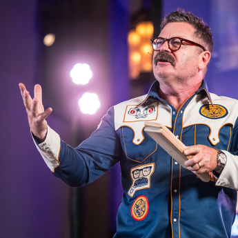Nick Offerman in a colorful shirt with patches speaks on stage, holding a wooden block with bright lights in the background.