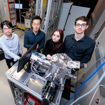 Four people in professional attire standing around advanced scientific equipment in a laboratory with computers and machinery.