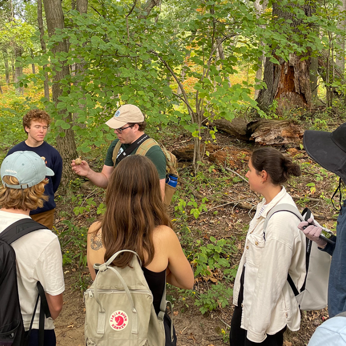 A group of students in backpacks and hats stand in a semi-circle in the woods. The all listen to a central speaker who is wearing a hat and holding a round natural object.
