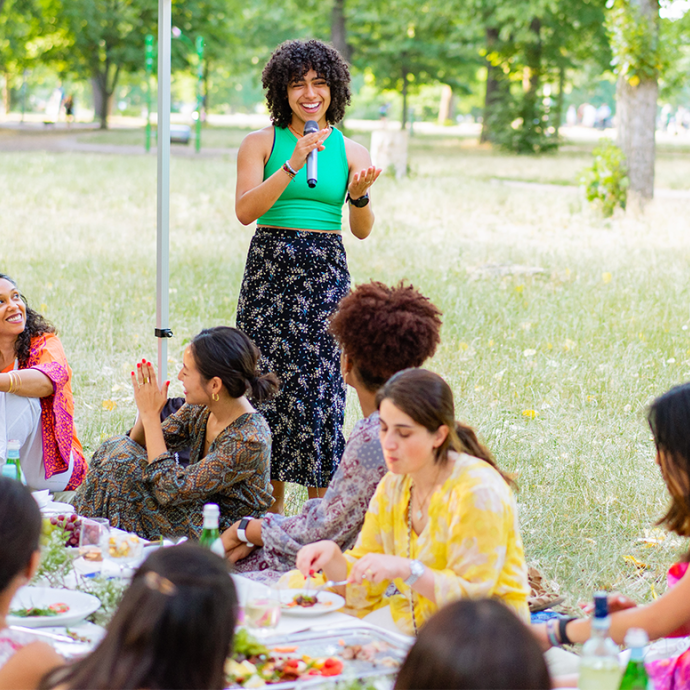 Dannerys Peralta stands holding and speaking into a microphone in front of a group of women sitting and eating at a long table outside.