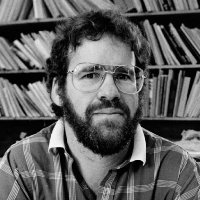 Black and white photo of a man with beard wearing glasses and looking at the camera in front of a wall of books