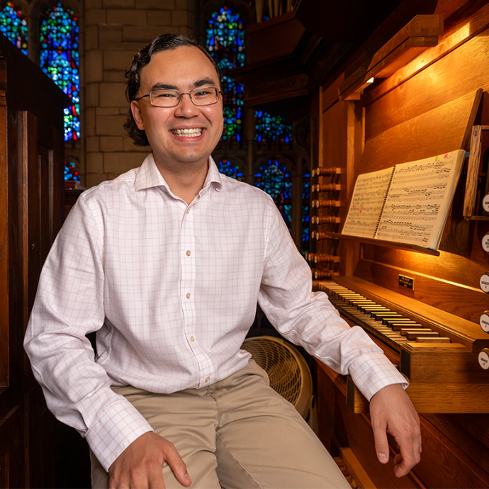 Jacob Reed sits on the bench next to an organ keyboard and stops. 