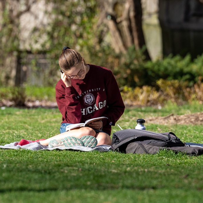Student wearing a University of Chicago sweatshirt reading a book while sitting on a blanket on the grass.